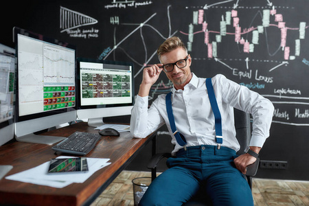 戴眼镜的中年白人交易员坐在电脑显示器前的办公桌旁看着摄像机.