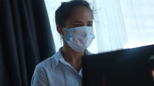 大流行期间被隔离在家的电脑前戴上口罩的妇女戴上医用口罩在笔记本上