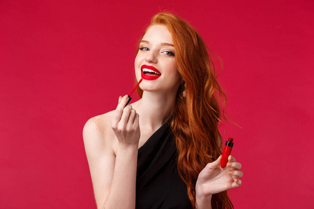 化妆美容和女性观念。肖像美丽的红发女人用唇膏红唇光泽和看着相机仿佛在镜子里，站在红色背景上。