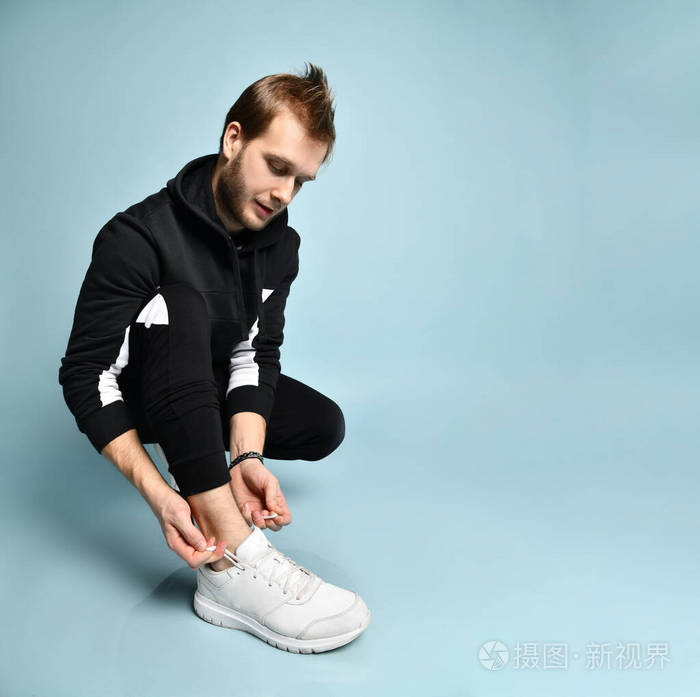 穿着黑色运动服手镯和运动鞋的时髦男人他蹲着系鞋带在蓝色背景上摆