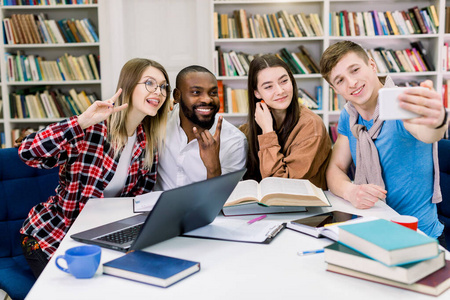 快乐的年轻大学生在图书馆学习时合影留念。大学图书馆里一群多种族的人坐在一起，拿着书和笔记本电脑