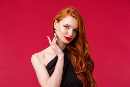用红色唇膏贴近性感迷人的红发女人的肖像，触摸耳环的相机，在派对上与人调情，穿着黑色礼服，红色背景，略带微笑。