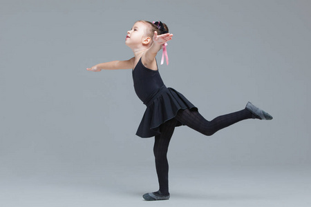 身着黑色运动服的漂亮小女孩将成为芭蕾舞演员，并在灰色背景上展示练习