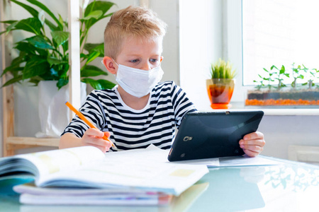 远程教育在线教育。病童戴着医用面罩在家学习，手里拿着数字平板电脑，做着学校作业。培训书和笔记本放在桌上。