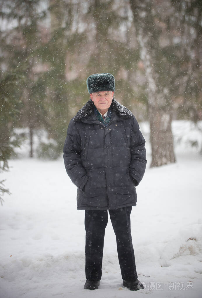 一位戴着温暖帽子的老人穿着羽绒服站在冬天的森林里照片-正版商用