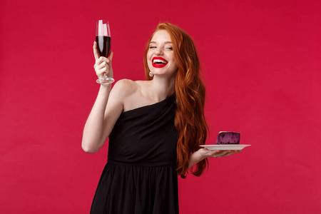 快乐华丽优雅的女性红头发女人举起香槟，说祝酒词或干杯，喝庆祝生日或在派对上玩得开心，穿着黑色连衣裙，红色背景