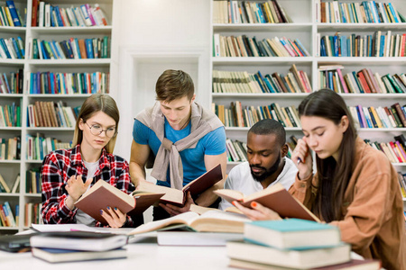 四个聪明而专注的多民族朋友坐在大学图书馆的桌子旁，一起看书，准备考试或上课，做家庭作业或搜索信息