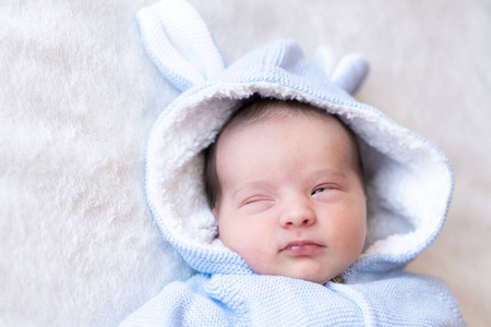一个熟睡的新生儿的特写肖像。在阳光明媚的托儿所里，一个穿着暖蓝色夹克和玩具兔耳朵的新生儿睡在毯子上。