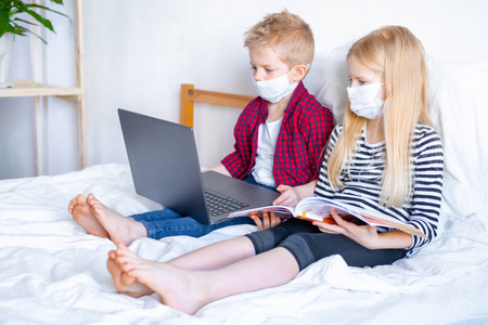 远程教育在线教育。疾病学校的男孩和女孩戴着医用面具在家里学习，用笔记本电脑和做作业。坐在床上拿着培训书