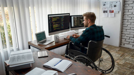 全神贯注。年轻的职业网络开发人员坐在轮椅上，坐在办公室里，在多个电脑屏幕上写程序代码