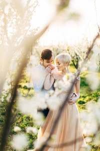 幸福浪漫的情侣在美丽的鲜花盛开的花园里散步。男人亲吻他漂亮女人的手。透过枝繁叶茂的枝头