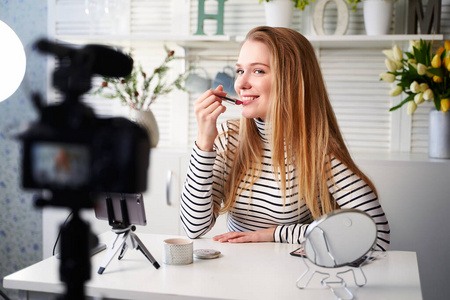 VLogg女性在唇部涂唇膏。美女博主女人在三脚架上用相机拍摄日常化妆教程。影响者金发女孩直播化妆品产品评论在家庭工作室