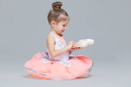 可爱可爱的芭蕾舞小女孩穿着粉色芭蕾舞裙坐在地板上，手里拿着芭蕾舞鞋