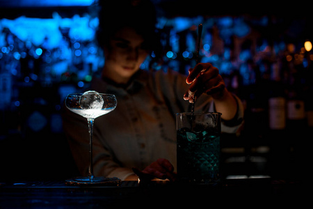 黑暗酒吧的女人用勺子在大玻璃杯里搅拌鸡尾酒。
