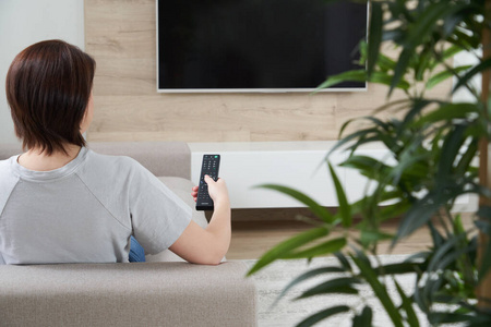 年轻女子坐在沙发上看电视,拿着遥控器照片