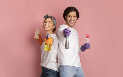 一对微笑的夫妇拿着喷雾清洁剂，站在工作室的粉红色背景上