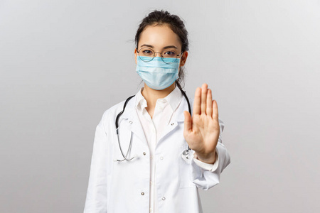 严肃的亚洲女医生戴着医用口罩,带着听诊器和白大褂,出示停止标志