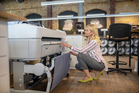 金发女人蹲在印刷机前，检查着什么