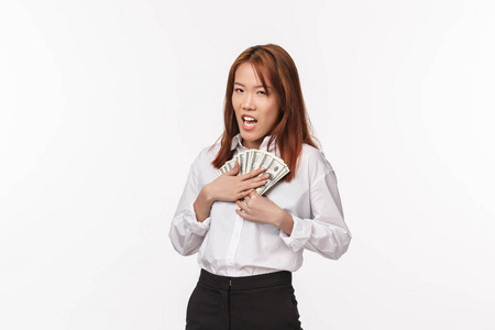 贪婪的年轻富有的亚洲女性身穿白衬衫,把钱压到胸前眯起眼睛怀疑,诡计