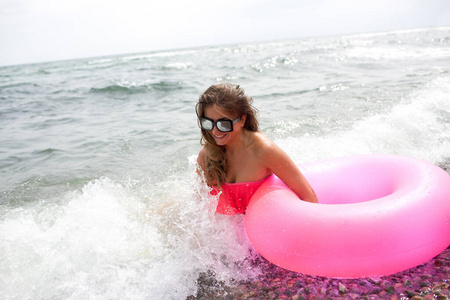 年轻女子坐在海边或海浪中，手里拿着粉红色的气垫，尽情享受。假期或旅行时间。图片上有鲜艳的彩色气垫和泳衣。