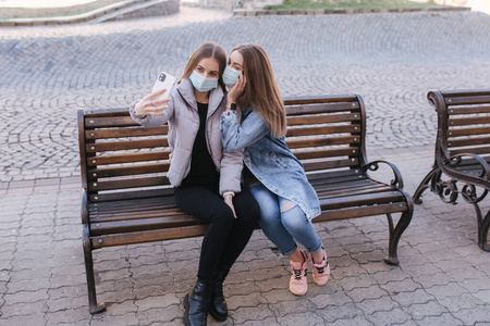 戴面具的女孩。冠状病毒主题。隔离期间，城里的妇女戴着防护面具坐在长凳上，用智能手机自拍