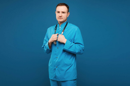 穿着蓝色医疗制服的帅哥医生拿着听诊器在蓝色背景上摆姿势。医疗保健和专业医学的概念