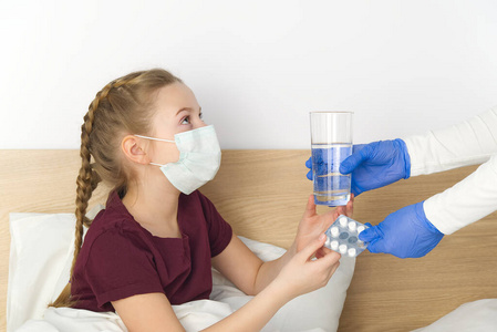 一个躺在床上的年轻女孩得到了药片和一杯水。蒙面女孩，防止病毒性疾病。病毒性疾病感冒和冠状病毒的检疫和治疗概念