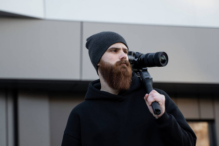 留着胡子的专业摄像师，穿着黑色连帽衫，手持专业摄像机，放在3轴万向节稳定器上。电影制作人用专业的电影摄影机制作一个很棒的视频。电