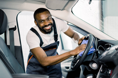 超细纤维和汽车内部清洁和细节。开朗微笑的非洲男性工人清洁室内现代汽车，汽车方向盘蓝色超细纤维衣服和清洁溶液