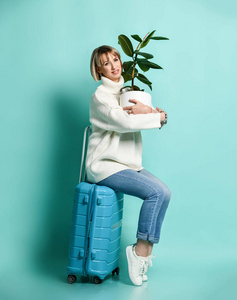 穿着白色毛衣牛仔裤和运动鞋的女人坐在一个蓝色的手提箱轮子上，手里拿着榕树，准备旅行