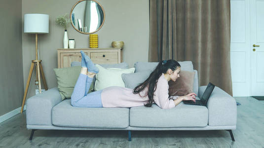 年轻的黑发女人躺在客厅的沙发上,在家工作,一边用笔记本电脑放松照片