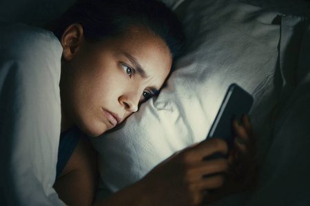 年轻漂亮的女人在床上用手机深夜在黑暗的卧室里,上网成瘾的概念照片