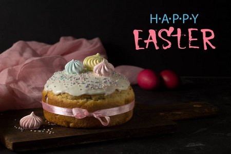 复活节蛋糕，黑色背景上有红色的鸡蛋和一块木制的砧板，一个漂亮的蛋白酥皮装饰。粉红色餐巾字母复活节快乐