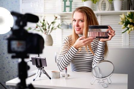 美女博主女人在三脚架上近镜头拍摄日常化妆教程。影响女孩直播化妆品产品评论在家庭工作室。Vlogger女性展示眼影调色板。