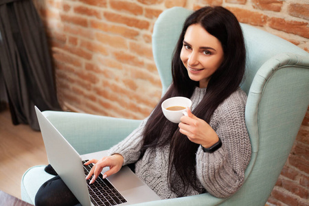 可爱的女人呆在家里，在笔记本电脑前喝咖啡