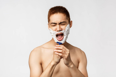 美人与卫生理念。无忧无虑的亚洲帅哥肖像刮胡子唱歌，在浴室里准备好，涂上面霜，对着剃须刀般的麦克风唱歌，站在白色的背景上
