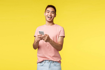 技术沟通和生活方式理念。热情的亚洲男性身穿粉色t恤，从酷酷的笑话或视频中笑出声来，用手机笑眯眯的相机，站在黄色背景下