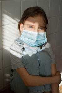 因为冠状病毒Covid19的流行和自我隔离，一个悲伤的小女孩正坐在厨房的白色瓷砖背景上，戴着一次性防护口罩