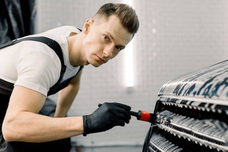 帅气的男性洗车工人看着摄像机，一边用一把特殊的刷子在一个汽车细节车间清洗汽车格栅。汽车服务业。