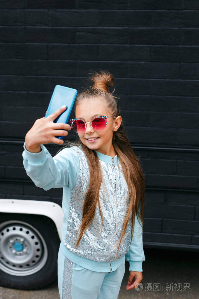 戴着墨镜的漂亮小女孩用智能手机自拍在户外微笑