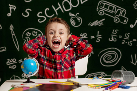 Emotional school boy sitting on the desk with many school suppli