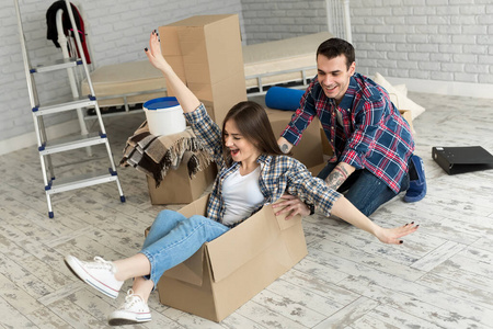 幸福的夫妇欢笑着搬进新家，年轻兴奋的女人坐在纸板箱里，男人推着纸板箱，兴高采烈的室友们一边玩耍一边收拾行李。