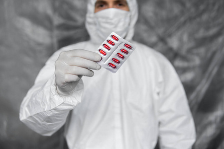 医生身穿白色防护服，戴着医用口罩和橡胶手套，手里拿着一种红色药丸，作为防止冠状病毒大流行的药物。流行，冠状病毒covid19大流