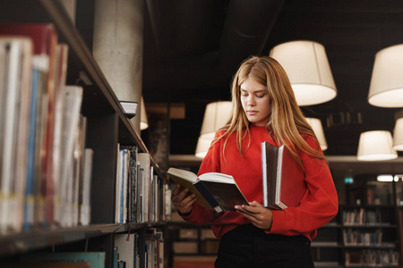 于校园图书馆里聪明的红发女孩侧视,站在书架旁,专注于看书,为下一堂