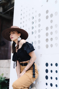 时尚的黑发女人，长发，戴着时髦的大柳条帽，对着一堵有洞的白墙摆姿势