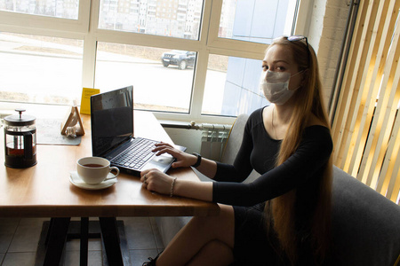 Beautiful girl sits and works at the table in a medical mask. On