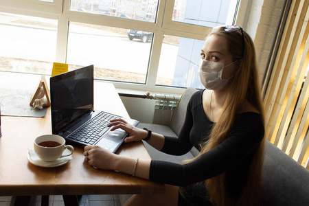 Beautiful girl sits and works at the table in a medical mask. On