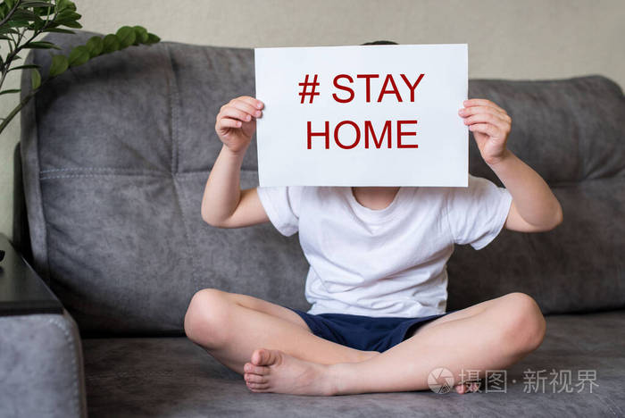 男孩坐在家里的沙发上拿着一张写着呆在家里的白纸捂住脸