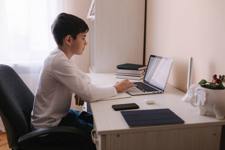 小学生在他房间的书桌旁学习。男孩用笔记本电脑上课。桌子上的书和写字板。在qurantine期间回家学习