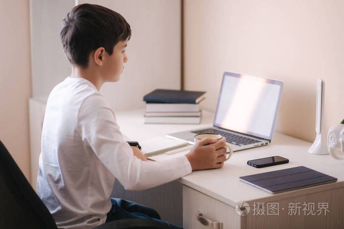 小学生在他房间的书桌旁学习男孩用笔记本电脑上课喝茶桌子上的书和